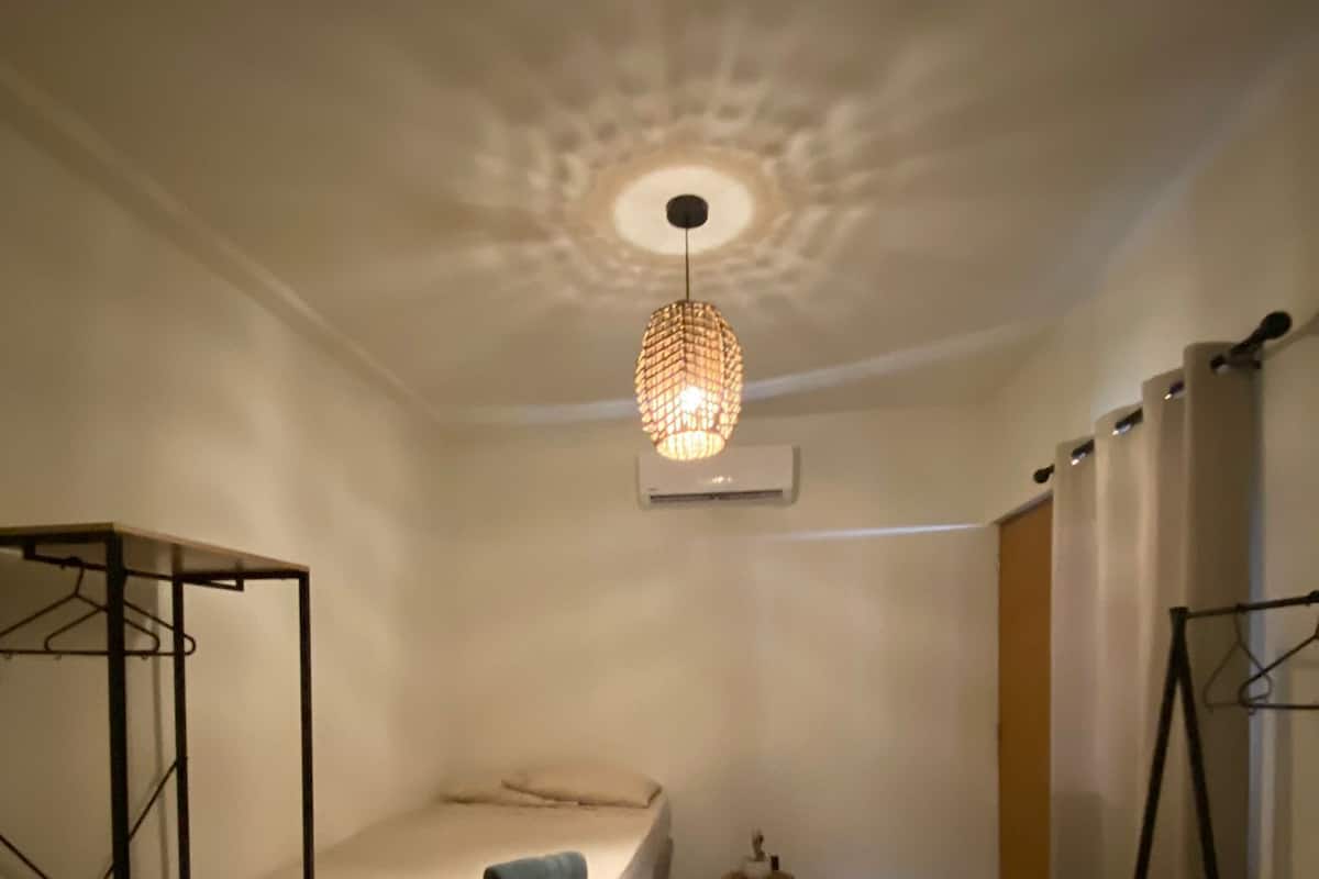 A small, well-lit bedroom with a single bed, a hanging light fixture, and a wardrobe.