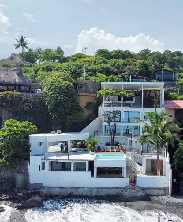 A multi-story beachfront property with a pool, balconies, and a thatched-roof structure nearby.