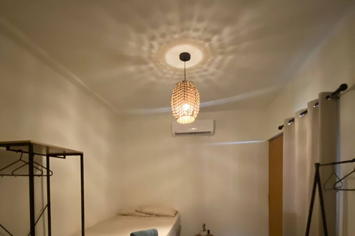 A small, well-lit bedroom with a single bed, a hanging light fixture, and a wardrobe.