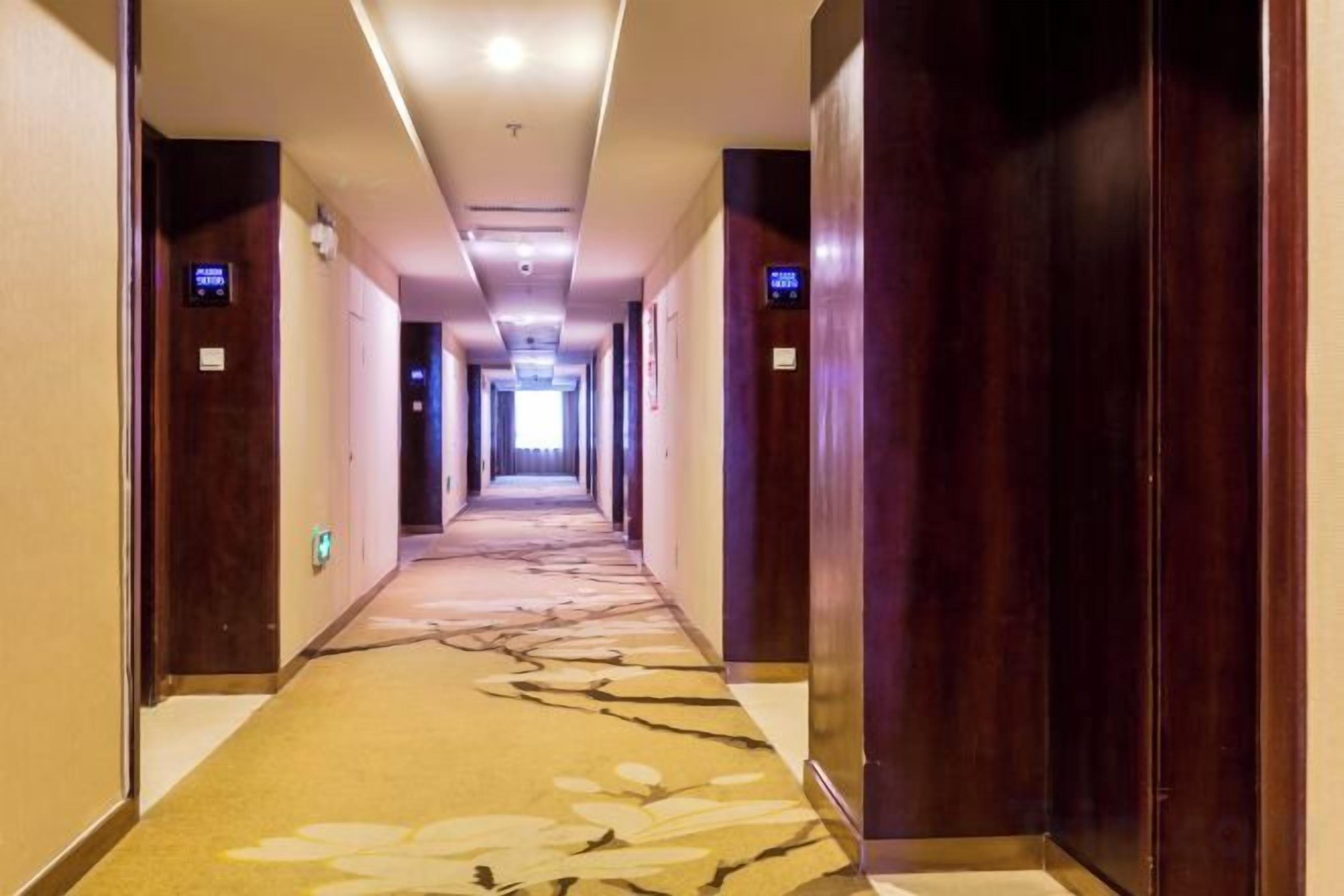 Pasillo de hotel con puertas de madera, suelo alfombrado y una fuente de luz al final.
