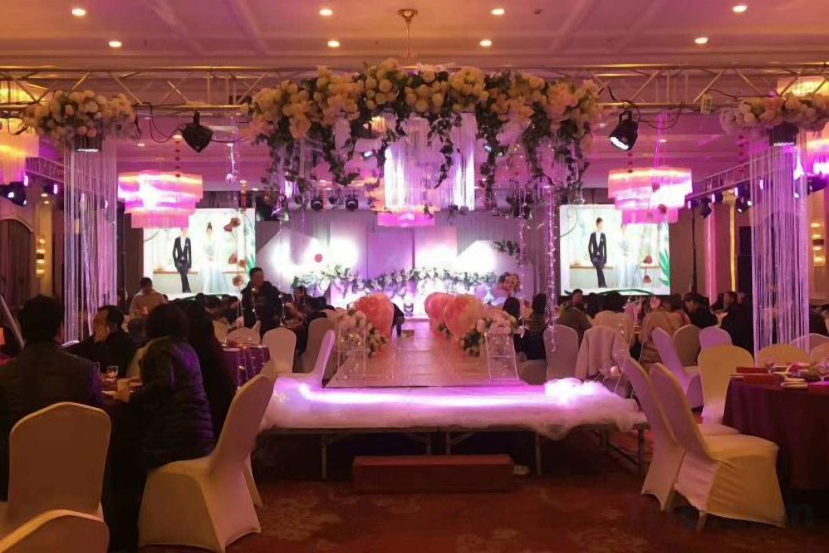 Un salón de banquetes con mesas redondas, sillas blancas y un escenario decorado con flores.