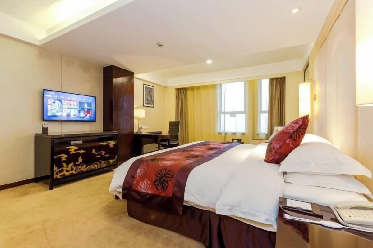 A hotel room with a bed, a desk, a television, and a large window with curtains.