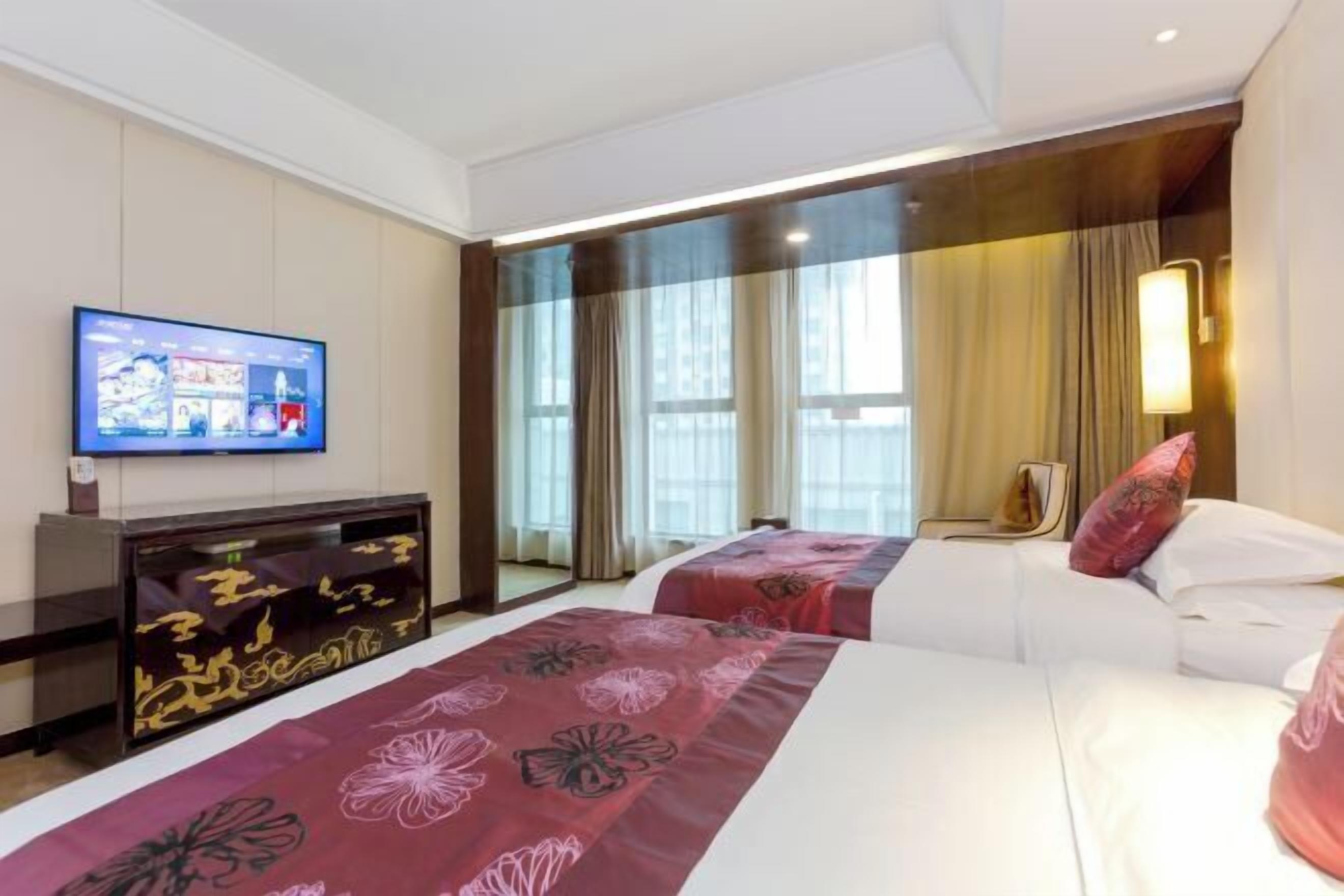 A hotel room with two beds, a TV, a desk, and a large window with curtains.
