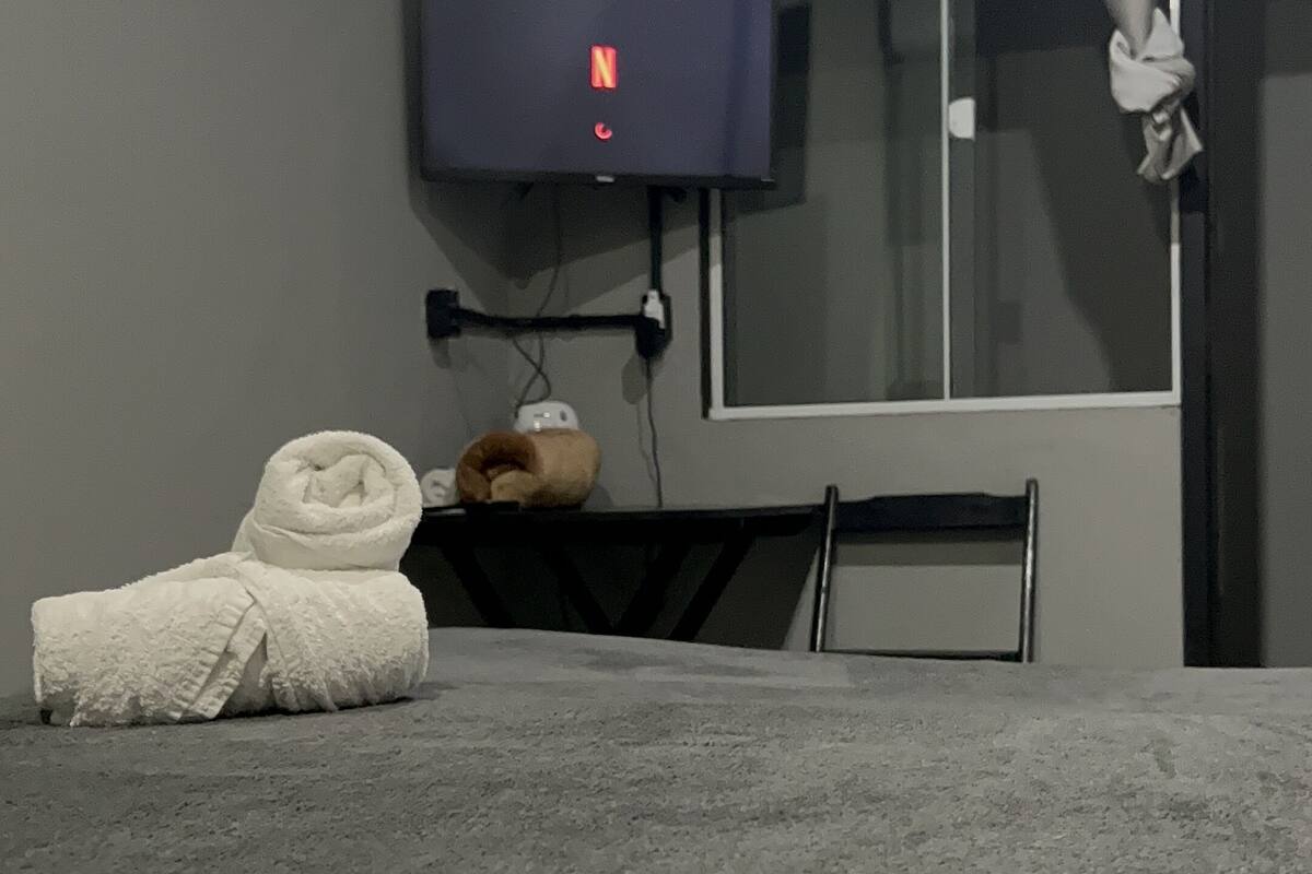 Ein Zimmer mit einem Bett, einem an der Wand befestigten Fernseher, einem Fenster mit Vorhängen und einem kleinen Tisch mit einem Handtuch und einem Stofftier.
