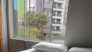 A hotel room with a bed, a red blanket, and a window with a view of buildings and trees. - Esquina 8 Hostel (Florianópolis)
