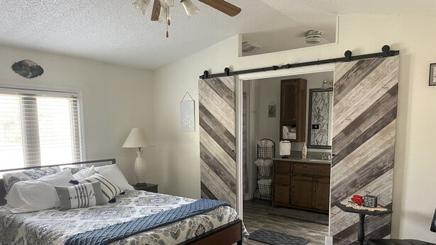 A bedroom with a bed, a nightstand, a lamp, a wooden floor, a bathroom with a mirror, a towel rack, and a bench.