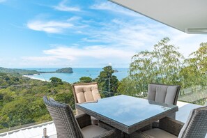 A balcony with wicker furniture, a glass table, and a view of a coastal landscape.