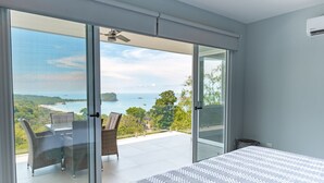 A bedroom with a large bed, a balcony with a table and chairs, and a view of the sea and greenery. - 🌅 Panoramic Ocean View Penthouse Suite at Villa Costa Vida (Quepos)