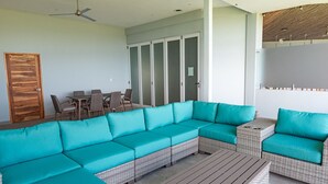A modern living room with a large teal sectional sofa, a wooden coffee table, wicker armchairs, and a dining set with chairs.