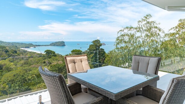 A balcony with wicker furniture, a glass table, and a view of a coastal landscape. - Villa&Tower -17 Bdrms,sleeps 55-AC,Elevator,Pool (Quepos)