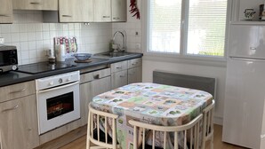 Une cuisine avec des meubles en bois, un réfrigérateur blanc, une table à manger recouverte d’une nappe à motifs de cactus et une fenêtre avec des stores.