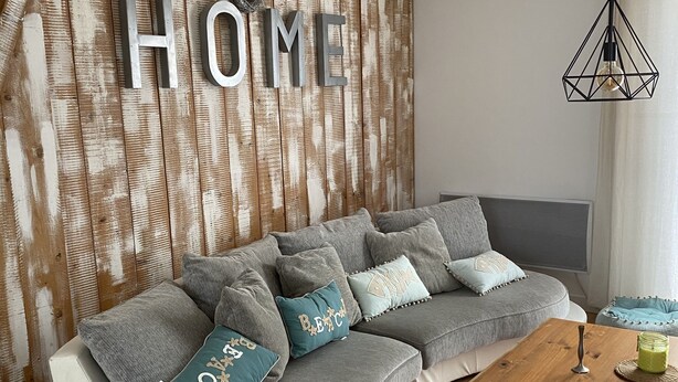 Un salon moderne avec un canapé gris, des coussins décoratifs, un mur d’accent en bois orné des lettres « HOME », une table basse en bois et un luminaire suspendu.