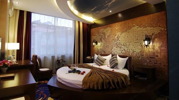 A hotel room with a large bed, a desk, a chair, a map on the wall, and a window with curtains. - Biancheng Hotel (Ruili)