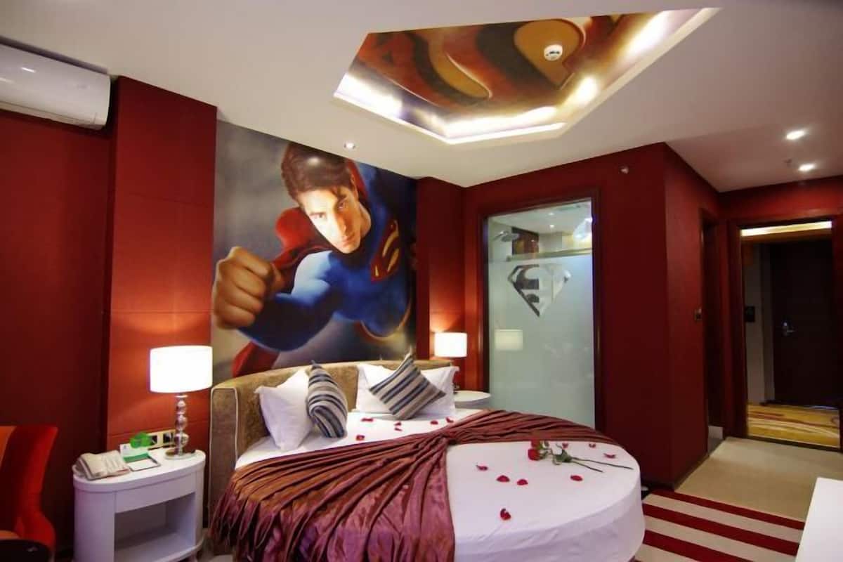A hotel room with a large superhero mural on the wall, a bed with a red blanket, and a nightstand with a lamp.