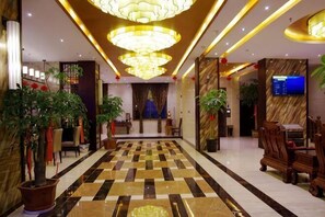 Um saguão de hotel com piso xadrez, lustres, plantas em vasos e um balcão de recepção.