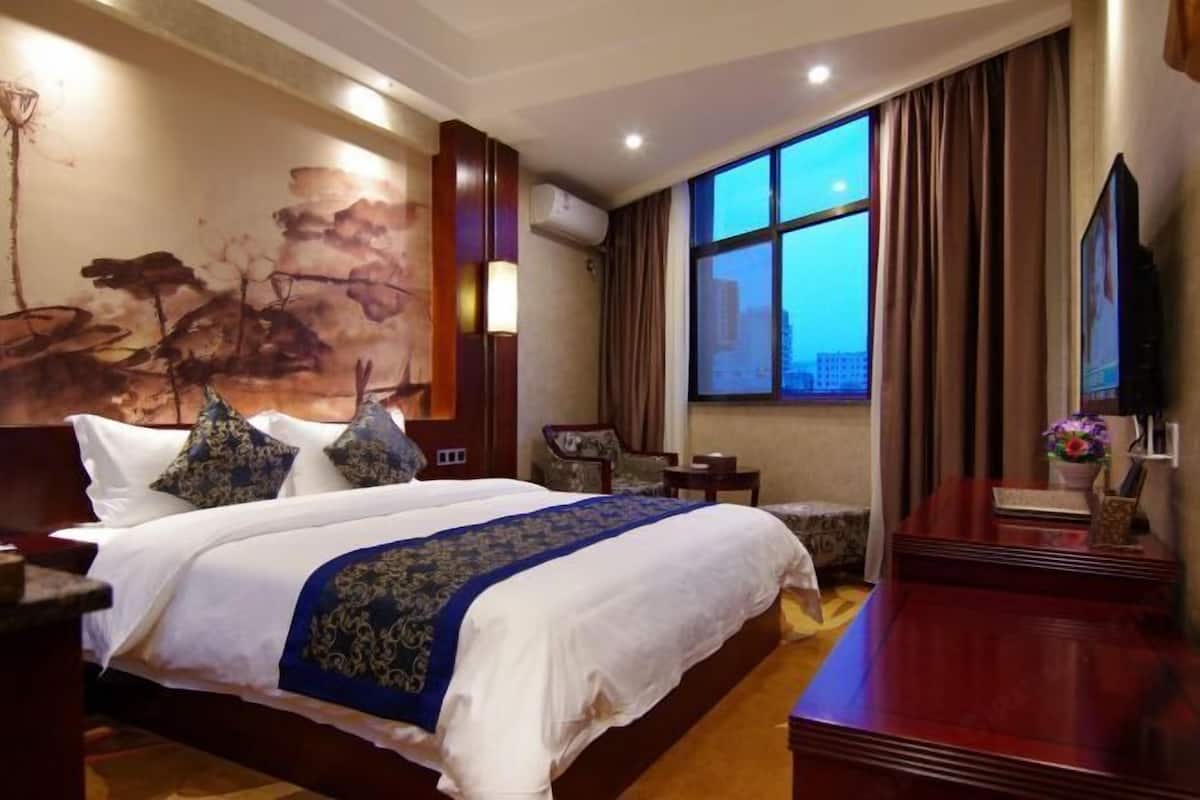 A hotel room with a large bed, a desk, a chair, a TV, and a window with a city view.