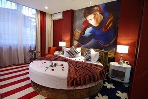 Quarto de hotel com uma cama grande, um tapete listrado em vermelho e branco, uma cortina vermelha, um mural na parede e uma pequena mesa de cabeceira com abajur.