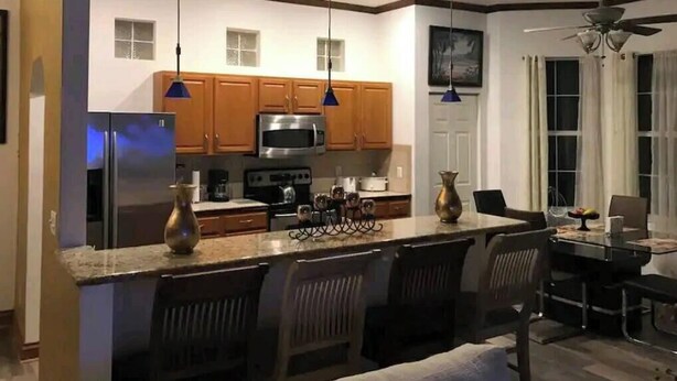 A modern kitchen with wooden cabinets, a refrigerator, and a microwave. There is a dining area with a table and chairs.
