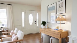 A well-lit living room with a wooden desk, a comfortable chair, a door with a glass panel, and framed artwork on the wall. - New! Refreshed modern coastal retreat in Duck! (Kitty Hawk)