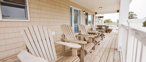 A covered porch with wooden Adirondack chairs and tables, a brick wall, and a white railing.