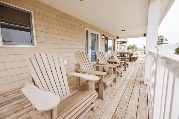 A covered porch with wooden Adirondack chairs and tables, a brick wall, and a white railing.
