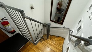Un escalier en bois avec une rampe métallique, un extincteur fixé au mur et un miroir qui reflète l’escalier.