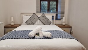 Un lit bien fait, avec une couverture à motifs, deux coussins décoratifs et une serviette enroulée posée sur une table de chevet.