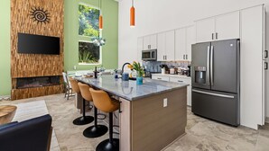 A modern kitchen with a central island, white cabinets, a large refrigerator, and a flat-screen TV mounted on a wooden wall. - 2 Homes 1 Incredible Stay*Private Pool & 5 Kings! (Panama City Beach)