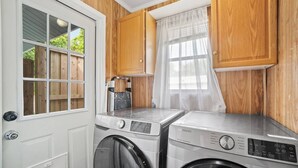 A laundry room with two washing machines, a drying rack, and a window with a sheer curtain.