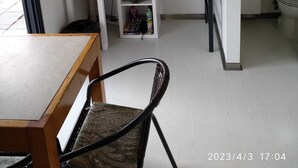 A compact room with a wooden table, a chair, a white shelving unit with various items, a television, and a bathroom visible through an open door.