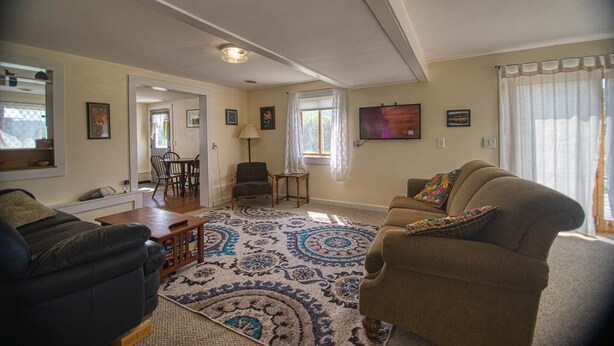 A living room with a sofa, armchairs, a coffee table, and a patterned rug. There is a television mounted on the wall and a window with curtains. - Cozy renovated barn apartment on first floor (Randolph Center)