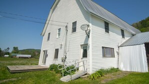 A white, single-story house with a gabled roof, a small porch, and a satellite dish. - Cozy renovated barn apartment on first floor (Randolph Center)