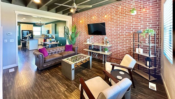 A modern living room with a sofa, coffee table, and chairs. There is a TV mounted on the brick wall and a kitchen in the background. - Charming updated LOFT style townhouse!!! (Midvale)