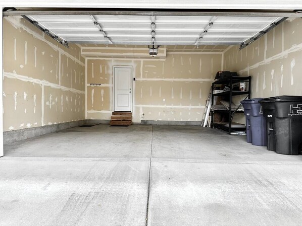 A garage with a concrete floor, a white door, and a metal garage door. There are two black trash bins and a black shelving unit against the wall.