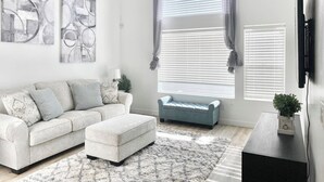 A living room with a sofa, ottoman, and a coffee table. There are abstract paintings on the wall, a window with curtains, and a flat-screen TV mounted on the wall.