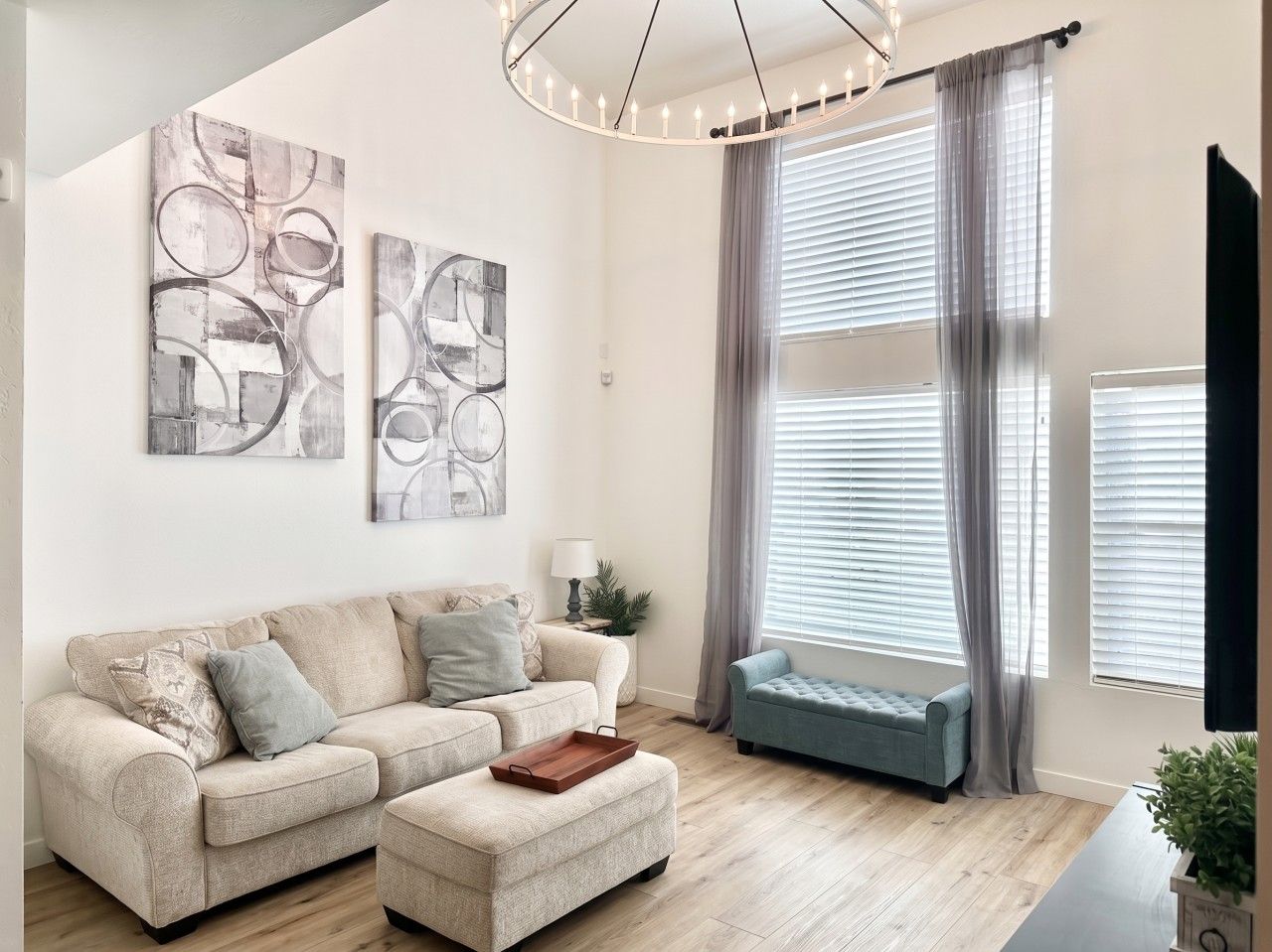 A living room with a sofa, ottoman, and a blue bench. There are abstract paintings on the wall and a lamp on a side table.