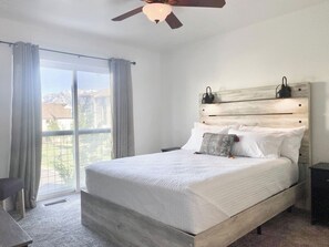 Ein Schlafzimmer mit Bett, Deckenventilator, Nachttisch und einem Fenster mit Vorhängen.