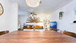 Uma sala de jantar com uma mesa de madeira, um sofá com almofadas, um ventilador de teto e uma televisão sobre um móvel.