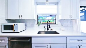 Uma cozinha moderna com armários brancos, uma pia de aço inoxidável, um micro-ondas e uma janela com vista para o verde.