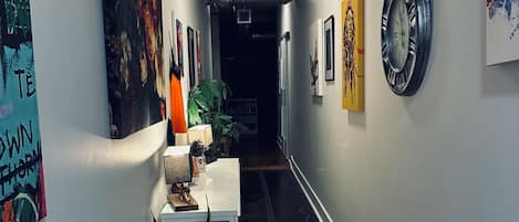 A hallway with artwork on the walls, a table with a lamp, and a checkered rug on the floor.