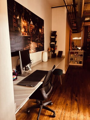 Un espace de bureau moderne comprenant un bureau, un ordinateur et un fauteuil de bureau. On y trouve une bibliothèque avec des livres et des objets décoratifs, ainsi qu’un grand tableau accroché au mur.
