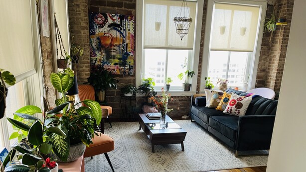 A cozy living room with a brick wall, wooden flooring, a black leather sofa, a coffee table, and various potted plants. - WorldCup Central- Easy access for sports and entertainment. (Houston)