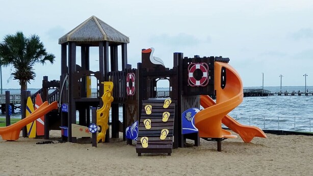 Une aire de jeux au bord de la plage, avec des toboggans et des structures d’escalade, située près d’une jetée et de palmiers.