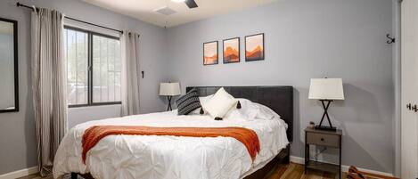 A bedroom with a bed, two bedside tables with lamps, a ceiling fan, a window with curtains, and a wall with three framed pictures.