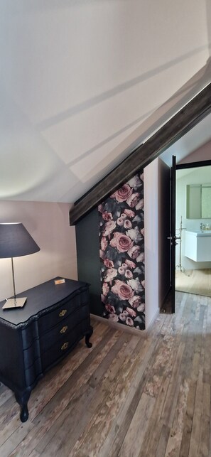 Una habitación con piso de madera, un aparador negro con cajones, una lámpara y papel tapiz con motivos florales en la pared.