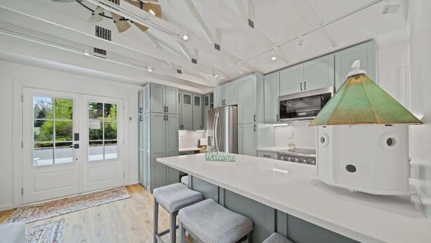 A modern kitchen with light blue cabinets, a white countertop, and a green pendant light. There are three stools and a rug on the floor. - Charming Downtown Cottage w/ Modern Amenities (Coeur d'Alene)