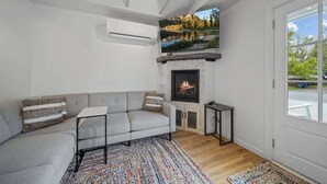 A modern living room with a fireplace, a gray sofa, a colorful rug, and a large window offering a scenic view. - Charming Downtown Cottage w/ Modern Amenities (Coeur d'Alene)