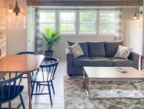 Ein gemütliches Wohnzimmer mit einem Holzespeisetisch, blauen Stühlen, einem grauen Sofa mit gemusterten Kissen und einem großen Fenster mit Vorhängen.