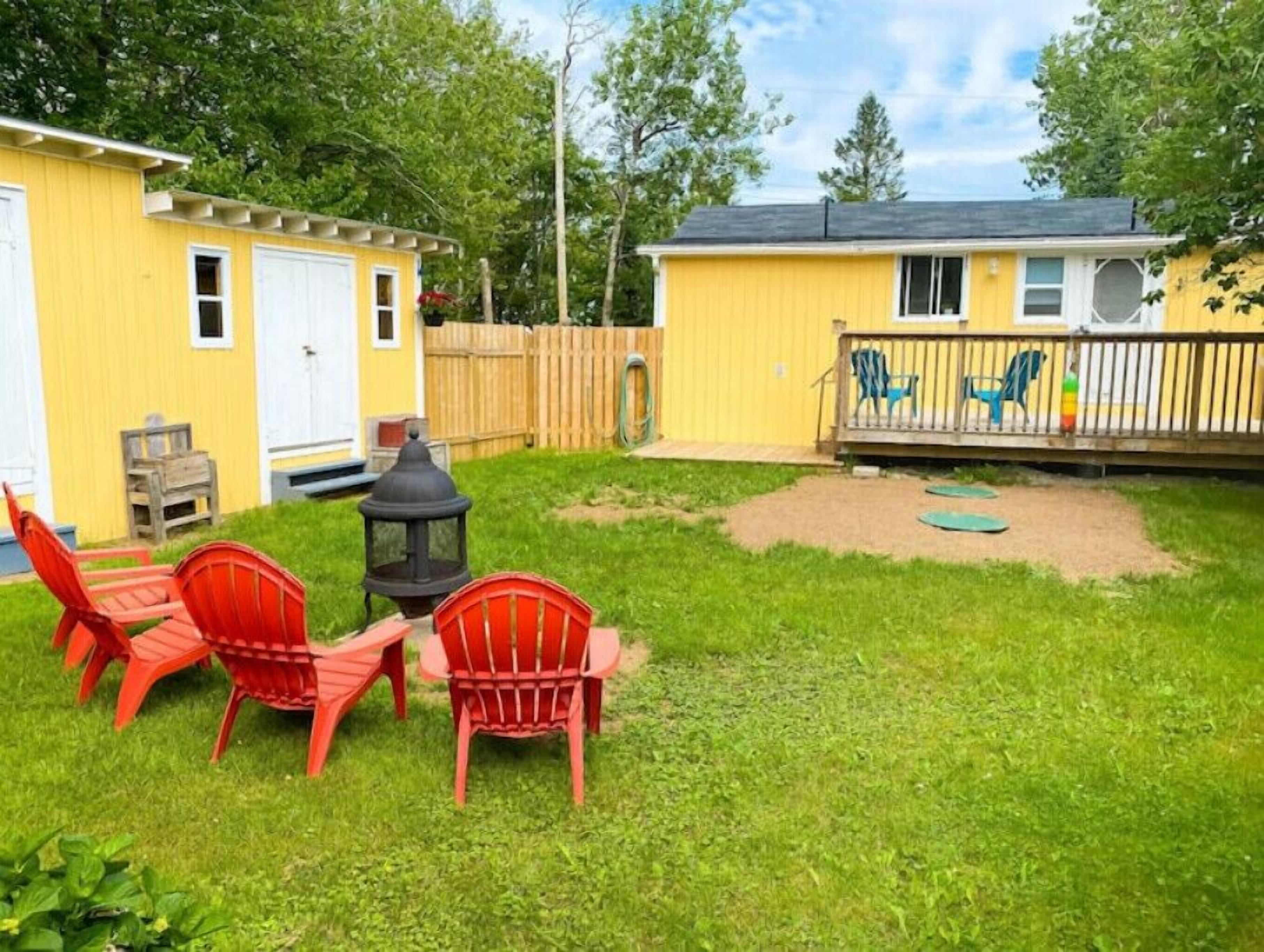Un jardin avec un abri de jardin jaune, deux chaises rouges, une lanterne et une terrasse en bois avec des chaises bleues.