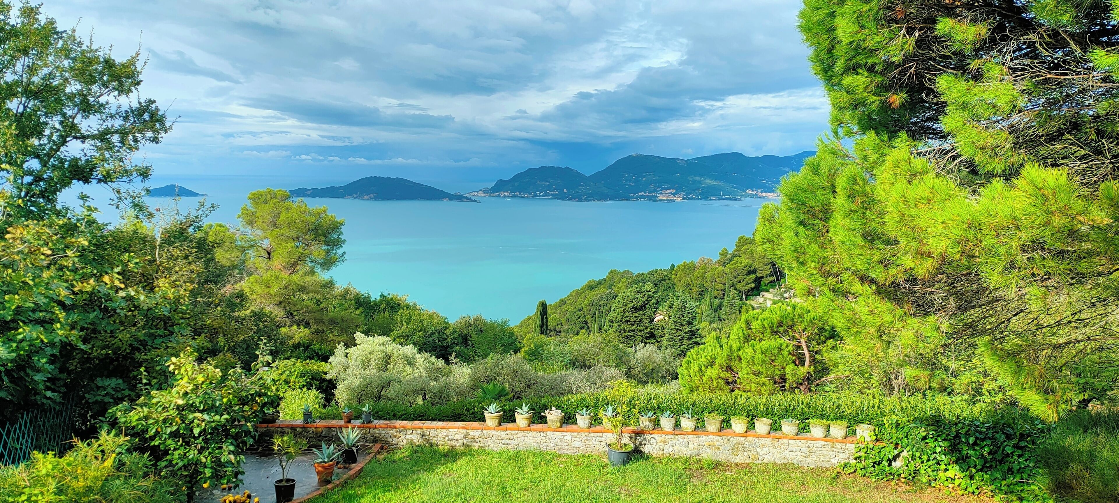 A lush garden with a stone wall and potted plants, overlooking a calm sea and distant mountains.